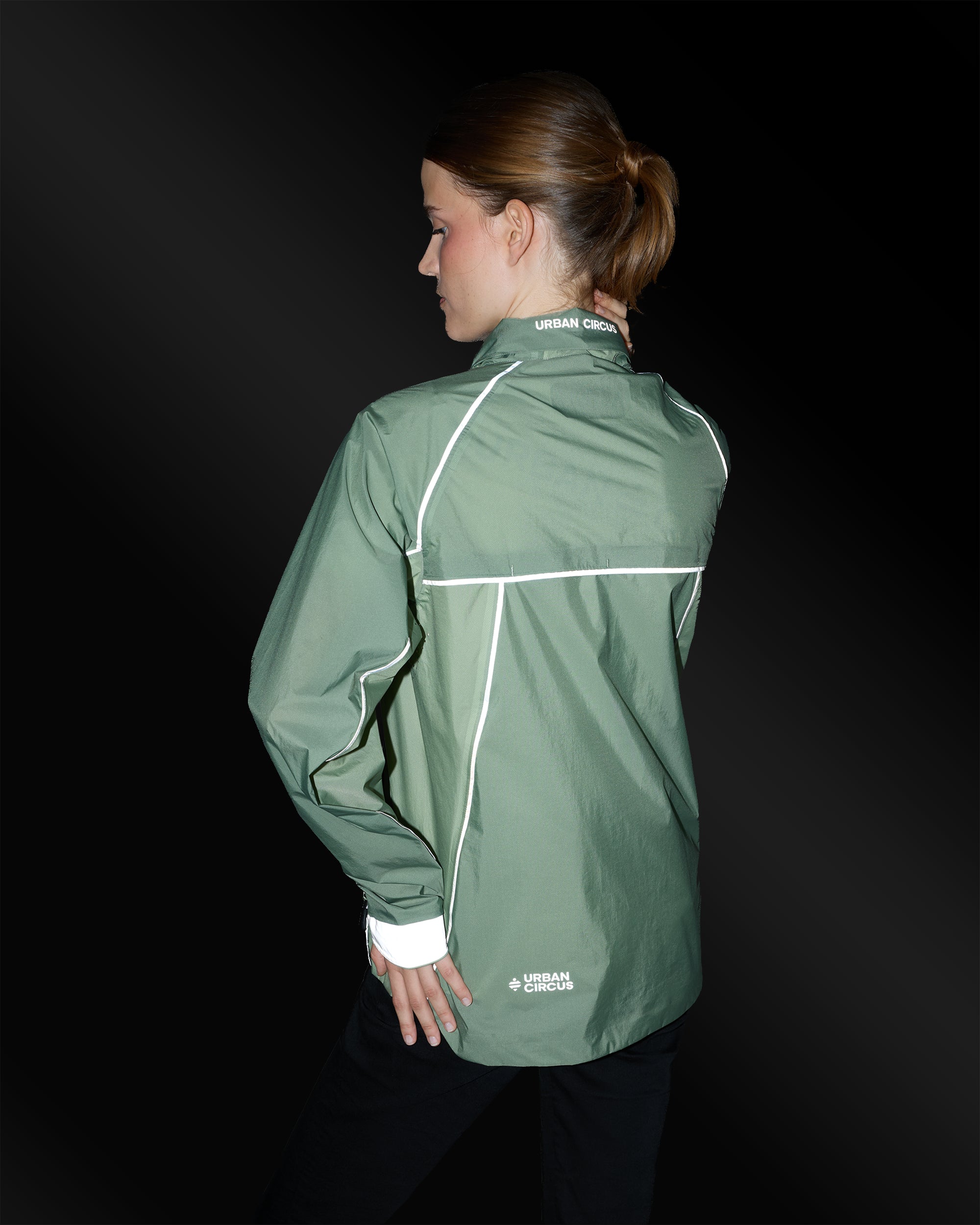 Compact ultralicht jack in saliegroen met reflecterende details, 's nachts gedragen door een vrouw van achteren